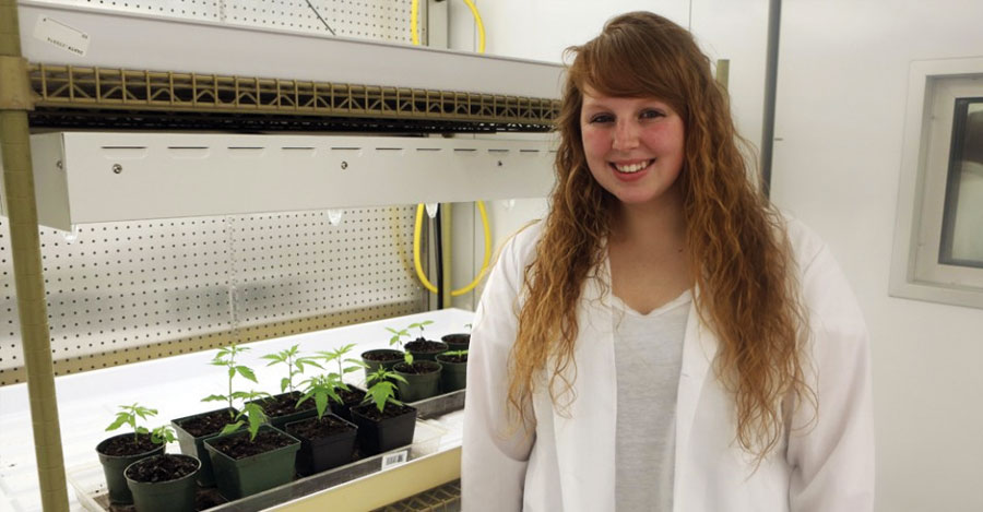 Cobleskill student Clara Richardson in lab coat in front of plants on table under light.
