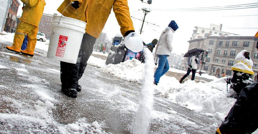 Man sprinkles salt on icey city sidewalk in winter.
