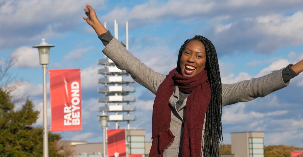 Stony Brook University senior Lydia Senatus stands outside on campus walkway with her arms stretched out to the sky.