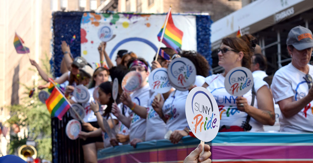 A parade float with SUNY Pride banner and people waving rainbow flags on it. 