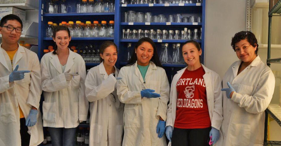 A girl in Cortland Red Dragons tshirt stands among others in white lab coats in a science lab.