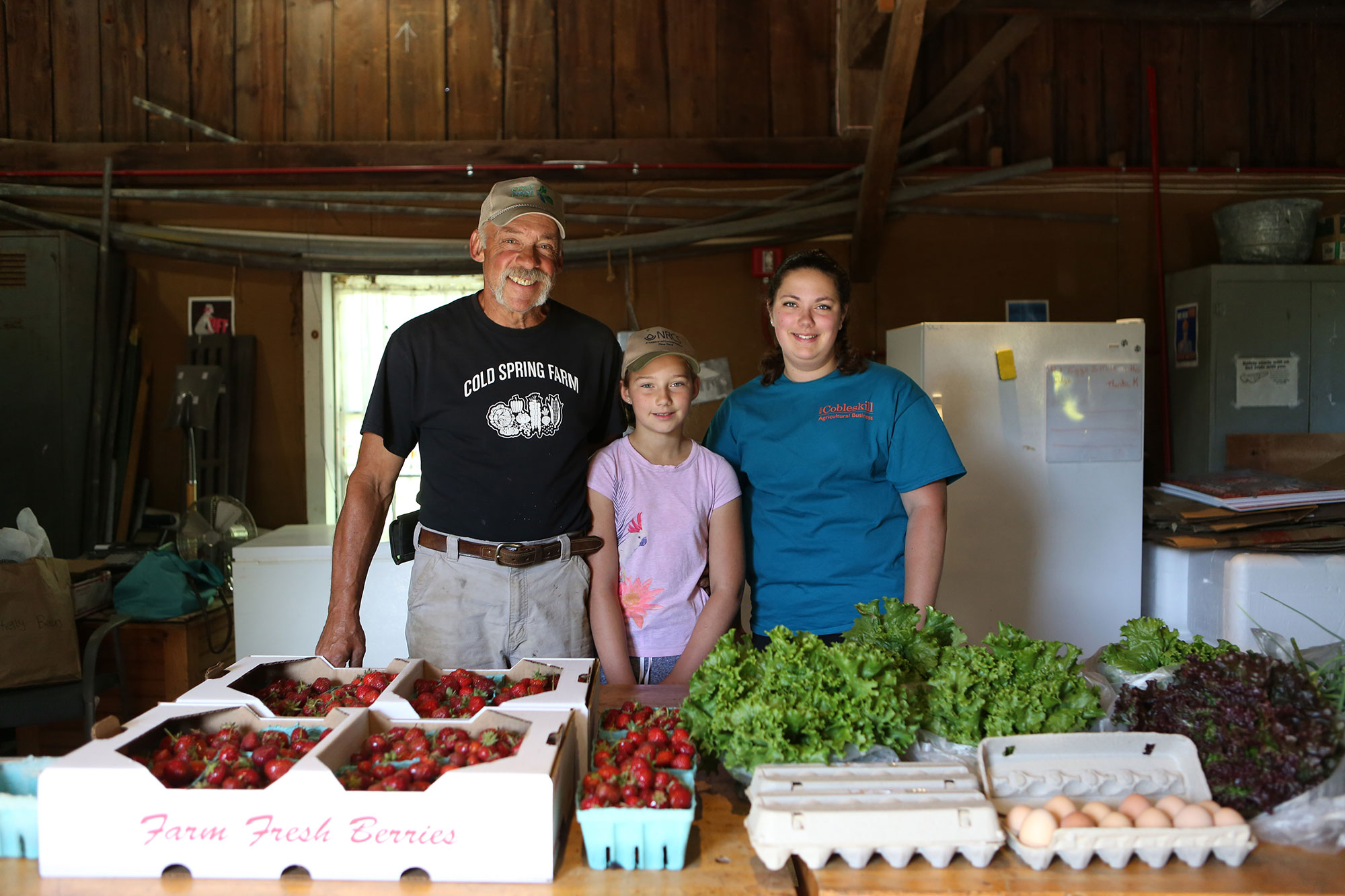 3 people stand behind a table of fresh produce and eggs being sold at the Scoharie Fresh General Store.