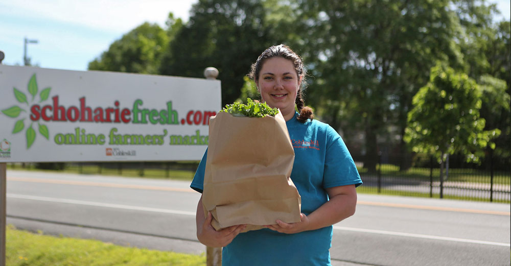 A female stands with a bag of fresh produce outside in front of the Scoharie Fresh store road sign.