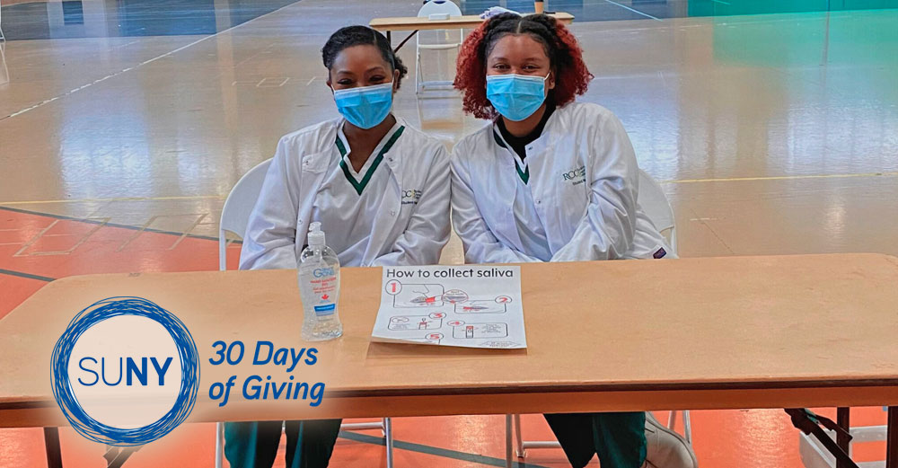 2 Rockland Community College students sit at a table in a gym wih face masks on, wearing nursing robe, ready to collect COVID-19 test samples.