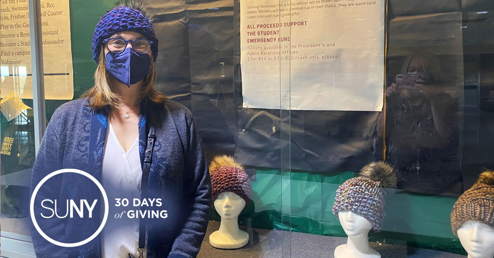 Female student stands in winter coat and face mask next to display of knitted hats.
