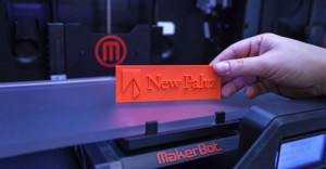 A 3D printed orange tag with SUNY New Paltz logo on it in front of MakerBot 3D printer.