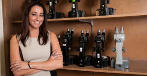 Michelle Miranda, associate professor at Farmingdale State College. stand in front of shelf of microscopes