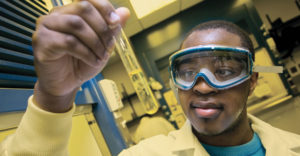Male students looks into beaker with solution in it while wearing goggles.