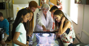 Teacher stands in dark room with students over x-ray in table.