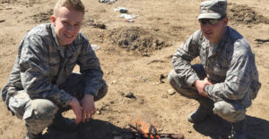 2 ROTC students from Farmingdale State College in uniform crouching down near self-made fire.
