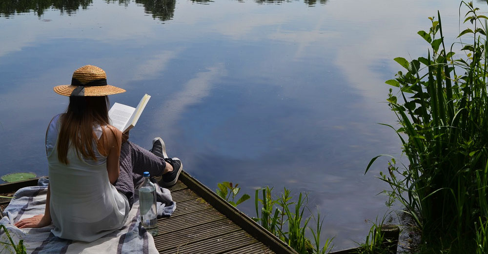 A girl reading a book on the dock overlooking a lake in the summer.