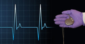 A goved hand holds a pacemarker over a heart beat chart.