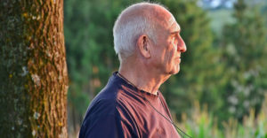 Male senior citizen stares to the right outdoors in front of trees.