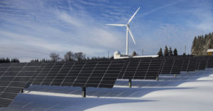 A windmill stands behind a field of solar panels in the winter snow.