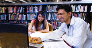 A male and female student review something on a laptop in the HVCC library.