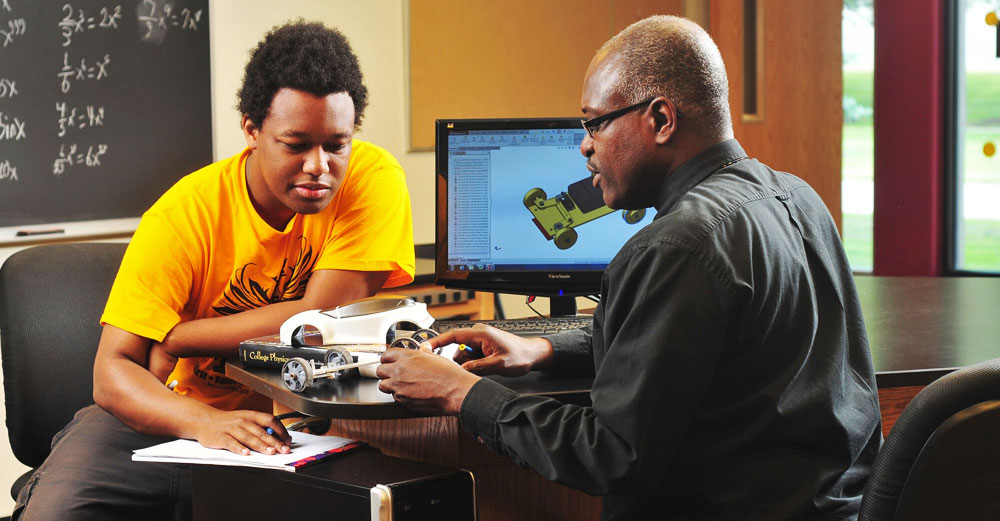 African-American engineering student sits with professor looking over model car.