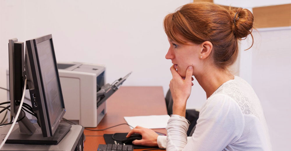 Red haired female student sits at a computer reading a website.
