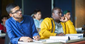 2 male minority students sit at desks in classroom listening to teacher.