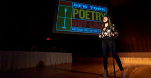 A girl stands on a wooden stage behind a microphone during a Poetry Out Loud competition.