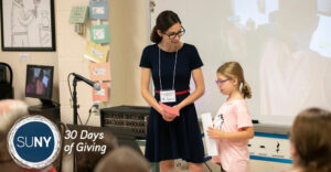 Female teacher stands next to elementary school student as she speaks in front of her class.