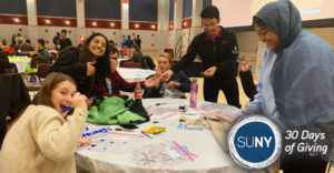Stony Brook University students smiling and waving while seated at a table full of art supplies in gym.