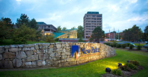 Stone entrance sign to SUNY New Paltz campus with lights on it during twilight hour.