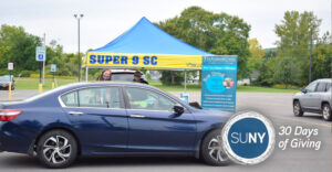 A blue car pulls up to a canopy tent in a parking lot at a socially distanced job fair.