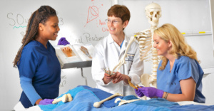 Two female students work with female teacher in a medical lab class looking at skeletons and bone structures.