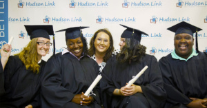 Diverse adult students smiling while seated in graduation cap and gowns and holding rolled up diplomas..