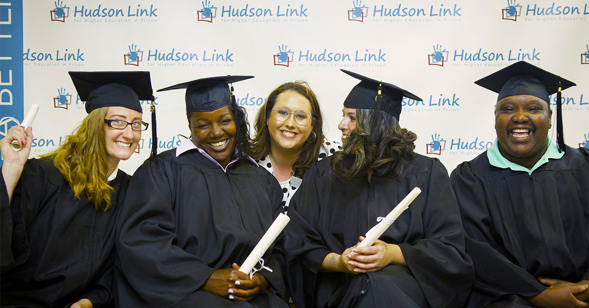 Diverse adult students smiling while seated in graduation cap and gowns and holding rolled up diplomas..