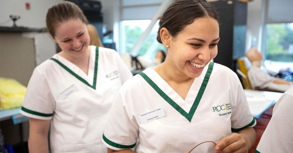 2 female nursing students in Rockland COmmunity College medical gowns smile in training lab.