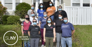 Students and employees from SUNY Upstate Medical University wearing facemasks and Upstate Strong t-shirts pose for a picture on steps in front of bungalow house.