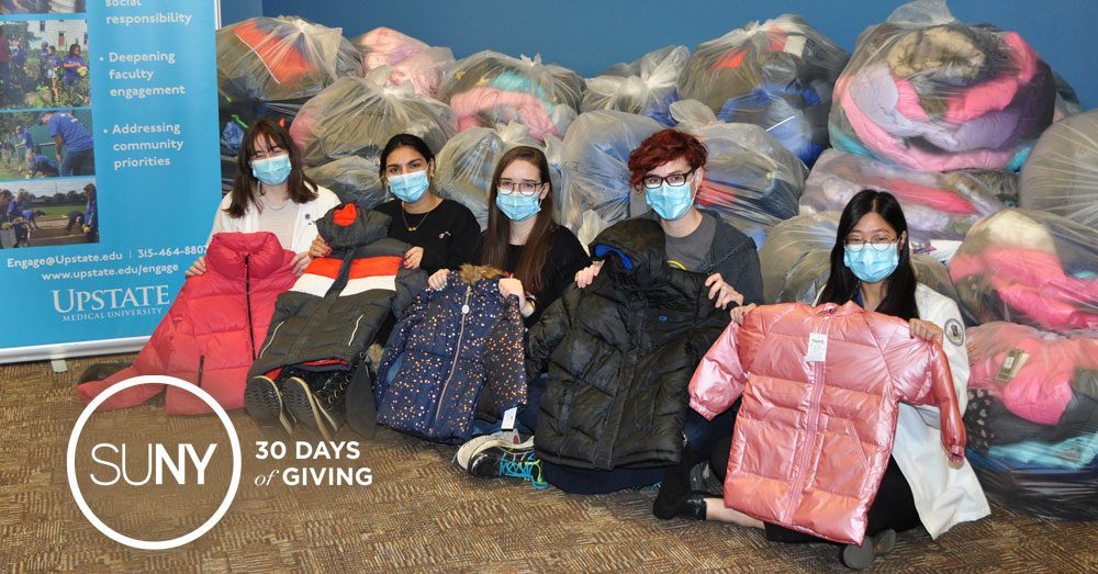 Students in face masks sit on floor in front of pile of bags filled with winter coats.
