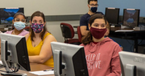Female college students sit in computer lab wearing face masks.