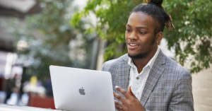 African American college student in a suit blazer sits in front of a laptop outside.