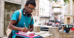 Male student sits on concrete ledge outside in city and writes notes in his textbooks.