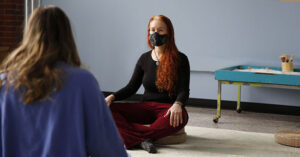 woman sits on floor with legs crossed preparing to lead meditation session.