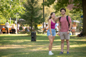 Two students pose together in large green space. People mill about the background.