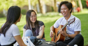 A male student smiles while plays guitar outside with two girls sitting around him.