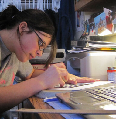 Girl writes at her desk in dorm room with laptop open.