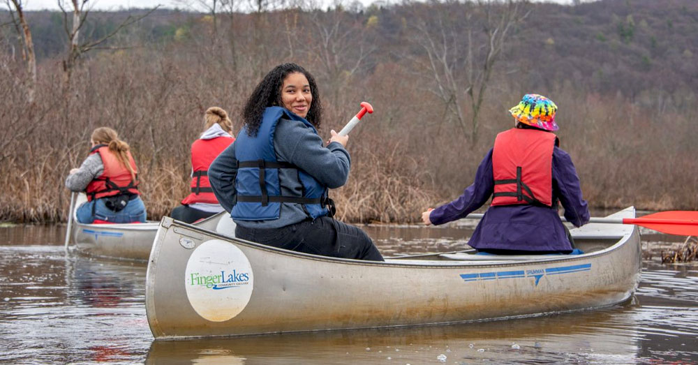 Girl paddls a canoe with Finger Lakes Community College logo on it.