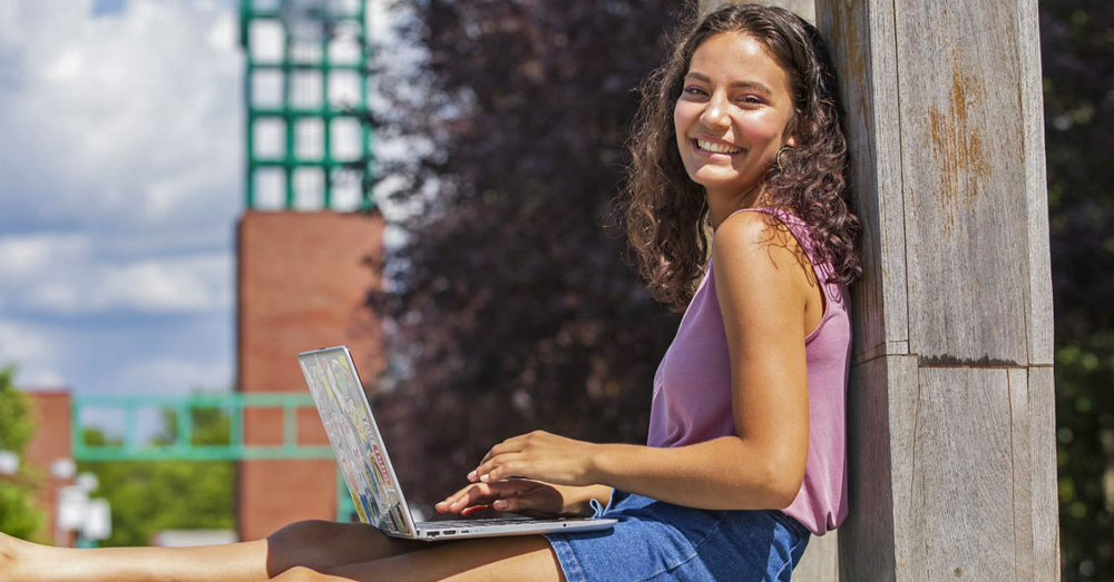 female student leans on wall outside with laptop in her lap on sunny day.