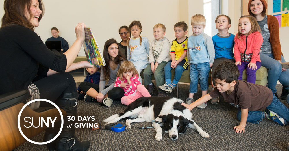 SUNY New Paltz dog Augie lays on a carpet with elementary school students while a teacher reads them a story.