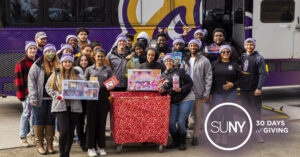 UAlbany students pose outside in front of a university school bus and a table full of gifts to be loaded onto it.
