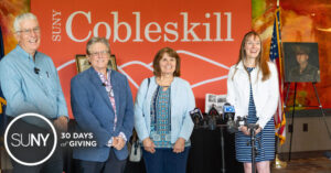 SUNY Cobleskill staff stand in front of school logo banner next to a painting of George Weitz.