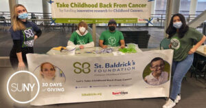 SUNY Oswego students holding a fundraiser at a table with a St Baldricks Foundation banner draped over it.