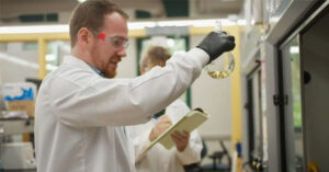 Dr. Christopher Thomasof SUNY ESF holds up a beaker in a lab while wearing protective white scrubs.