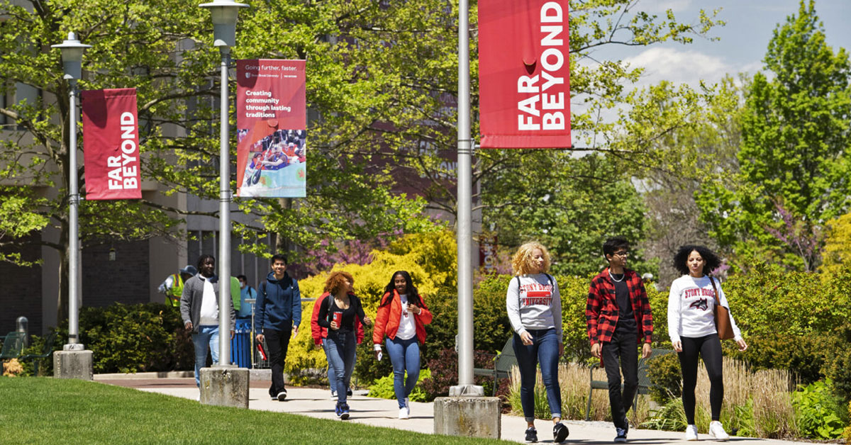Students walk along a tree lined sidewalk at Stony Brook University with banners hanging on light poles with Far Beyond printed on them.
