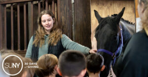 Female teacher holds a horse by his reigns in a stable barn to show elementary school students.