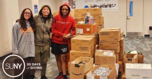 3 female student lacrosse players at Purchase College stand in a room with boxes of donated food stuff.